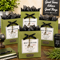 Four green Best Team Ever appreciation gift bags arranged in a professional office setting, featuring black ribbon, decorative key accents and separate custom company branding plates beneath the main labels.
